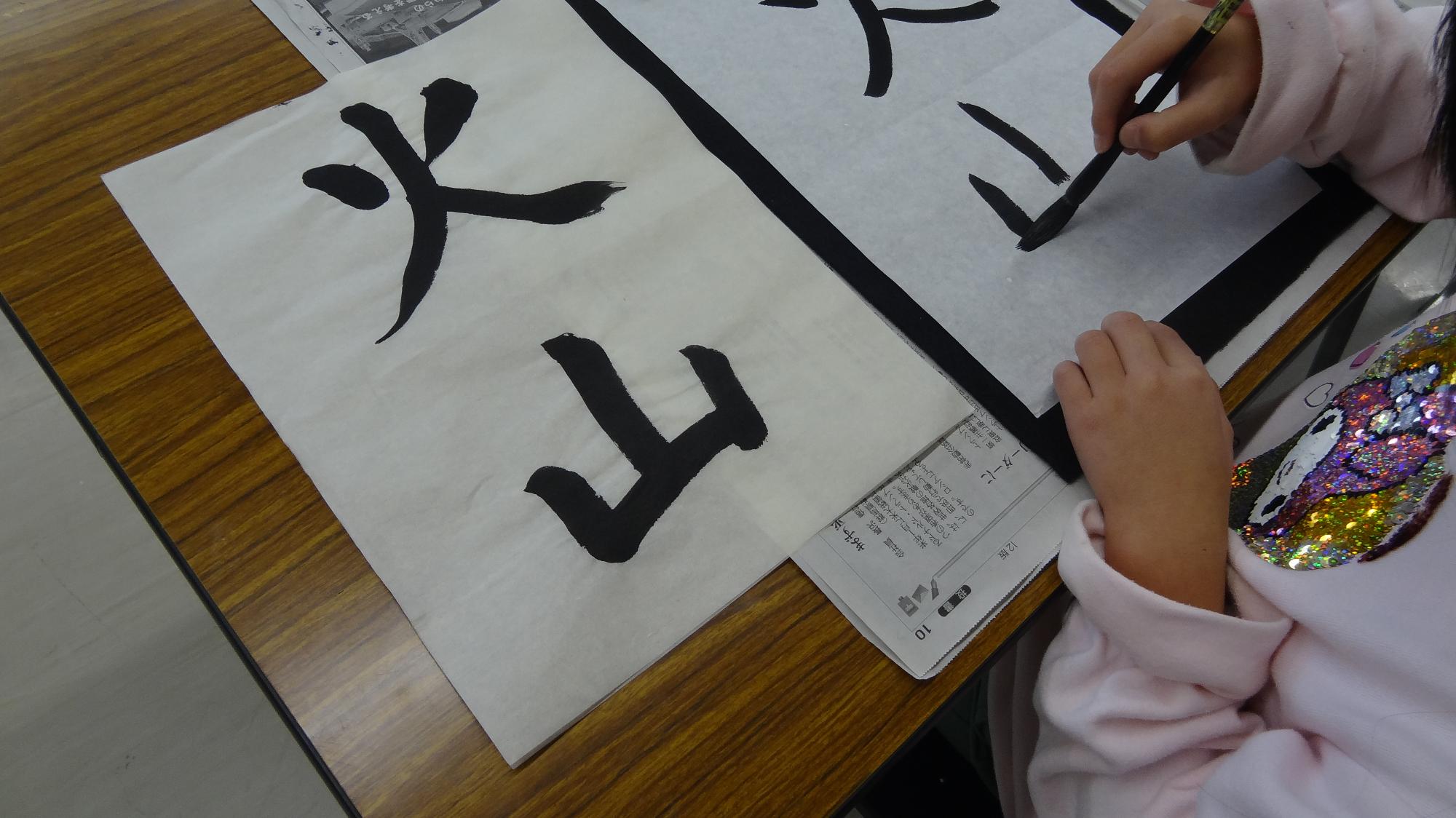 書道の行事で、見本見ながら、火山という漢字を書いている様子の画像