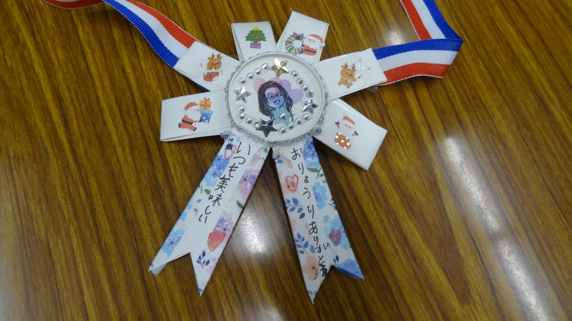 紙コップで作成したメダル。真ん中にお母さんの似顔絵が書かれていて、周りはシールが貼られている画像。いつも美味しいおりょうりありがとう、と書かれている