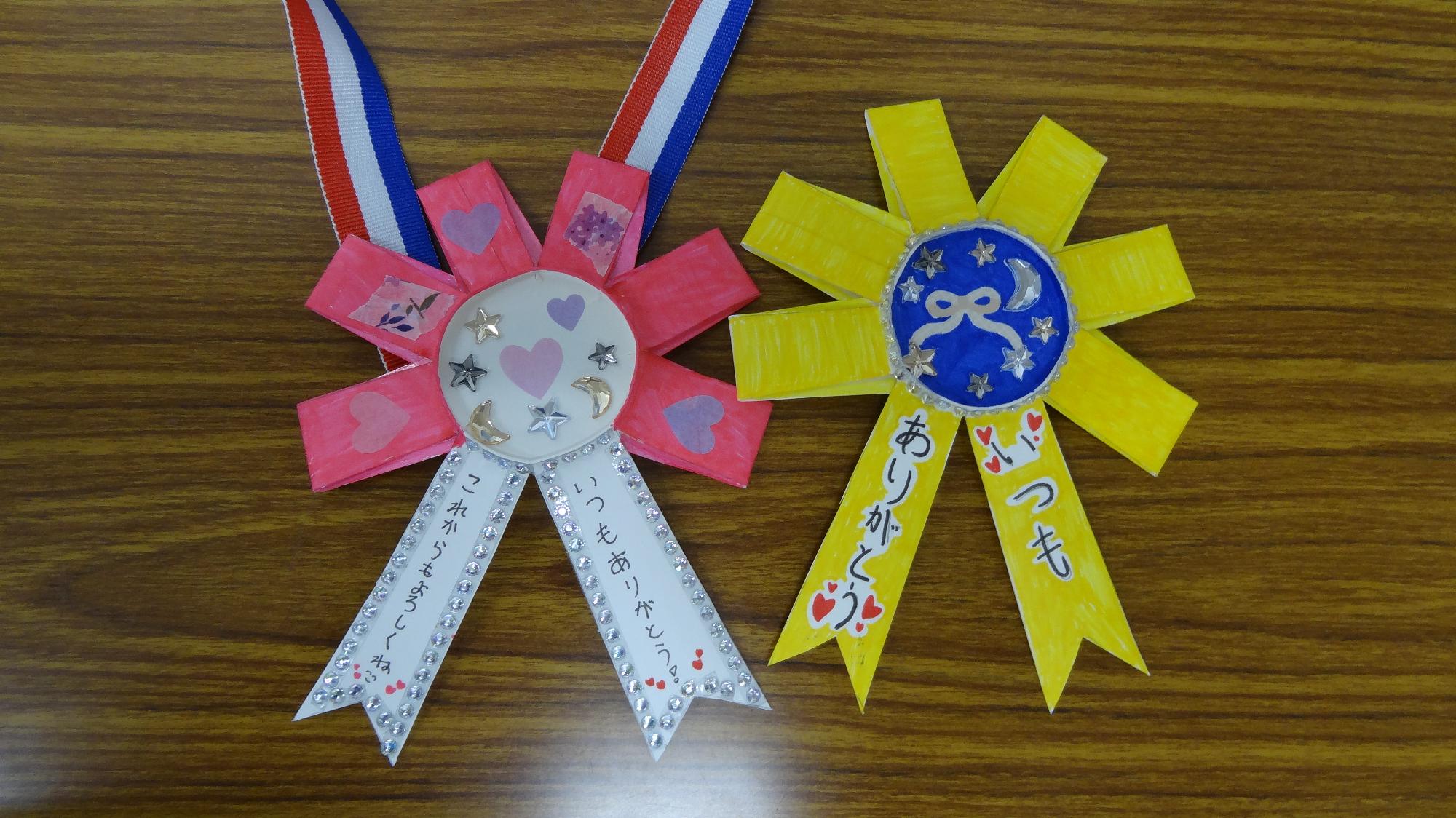 紙コップで作成したメダルが二つ並んでいる画像。左のメダルは周りがピンクで塗られていて、いつもありがとう、これからもよろしくねと書かれている。右のメダルは周りが黄色で塗られていて、いつもありがとう。と書かれている。