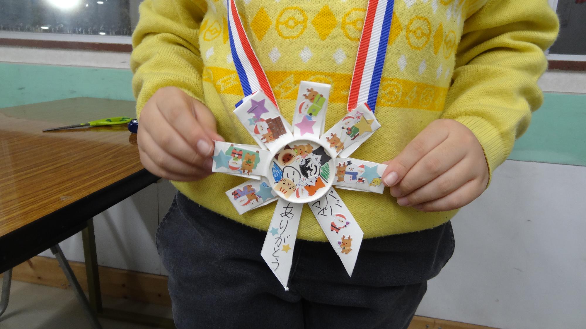 紙コップで作成したメダルを首にかけた児童の画像。メダルの周りはシールが貼られていて、真ん中には、人のイラストが描かれている。