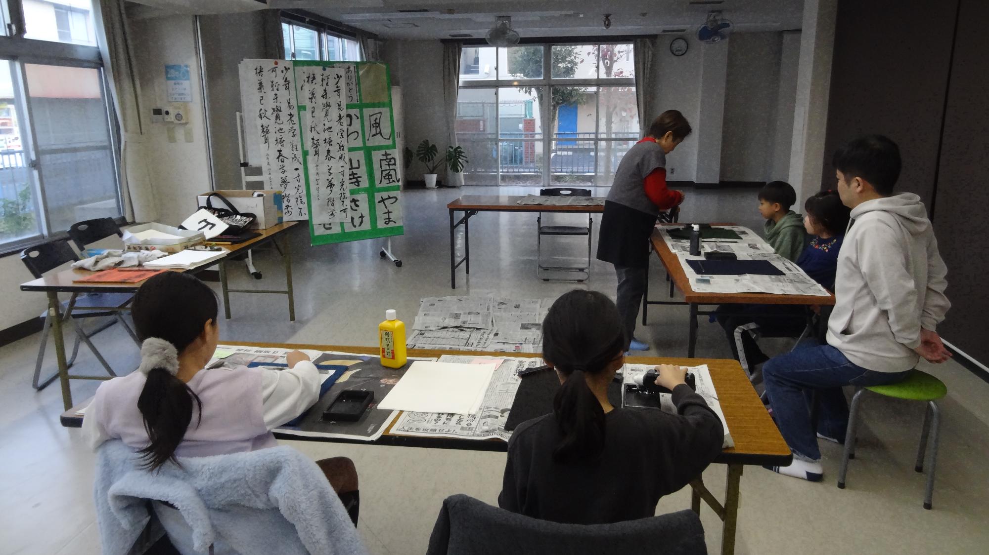 小学生4名が書道の講師1名に注目している様子。右側に1名の保護者が見守っている。