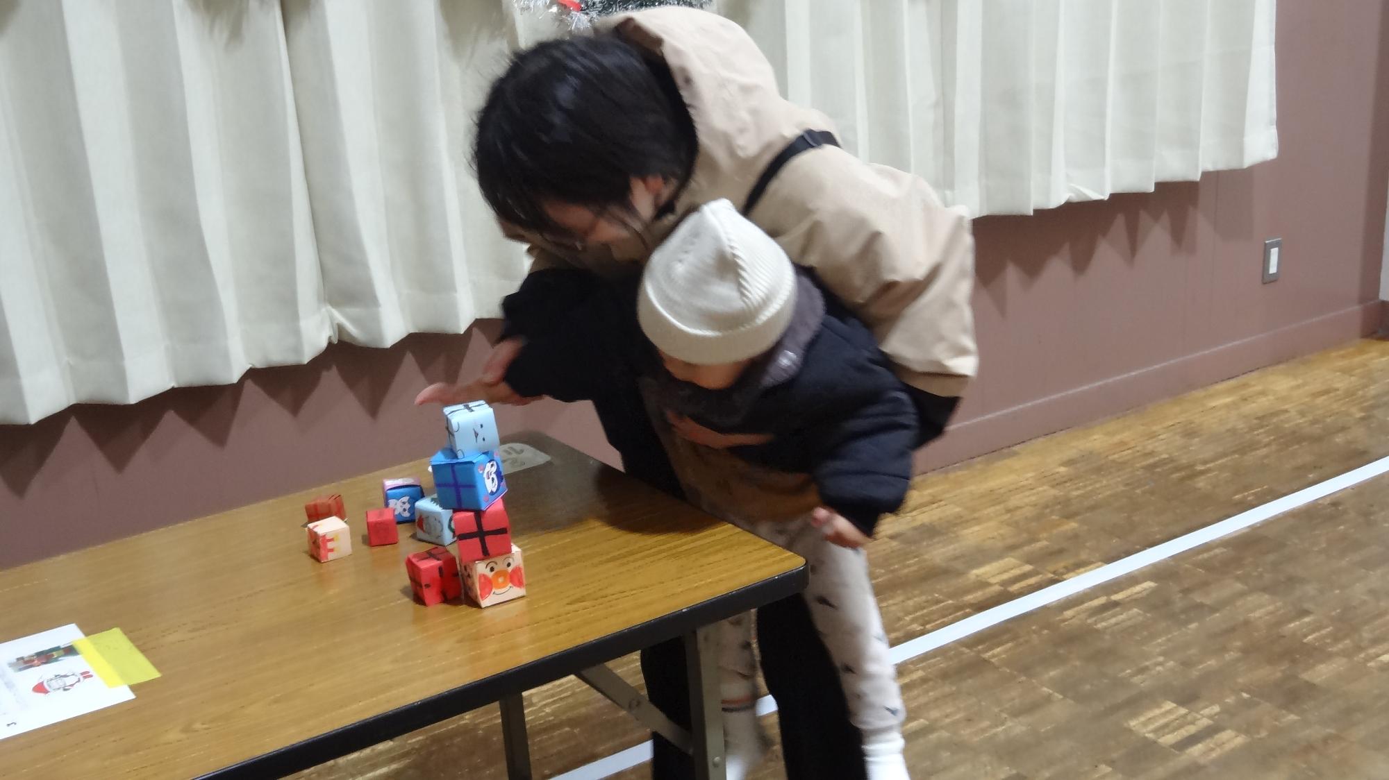 保護者にだっこされた幼児が折り紙でできたプレゼントを積んでいる様子。