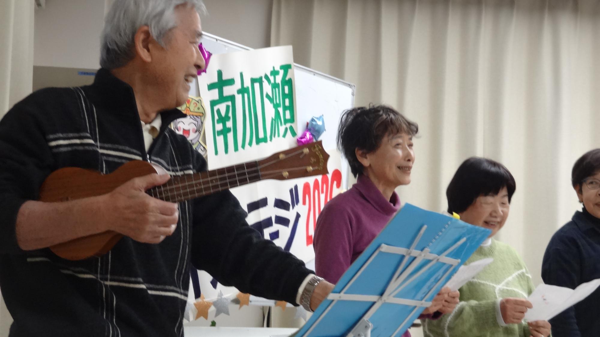 ウクレレを持った男性がにこやかに笑っている様子。その横には、笑顔な女性2名が映っている。