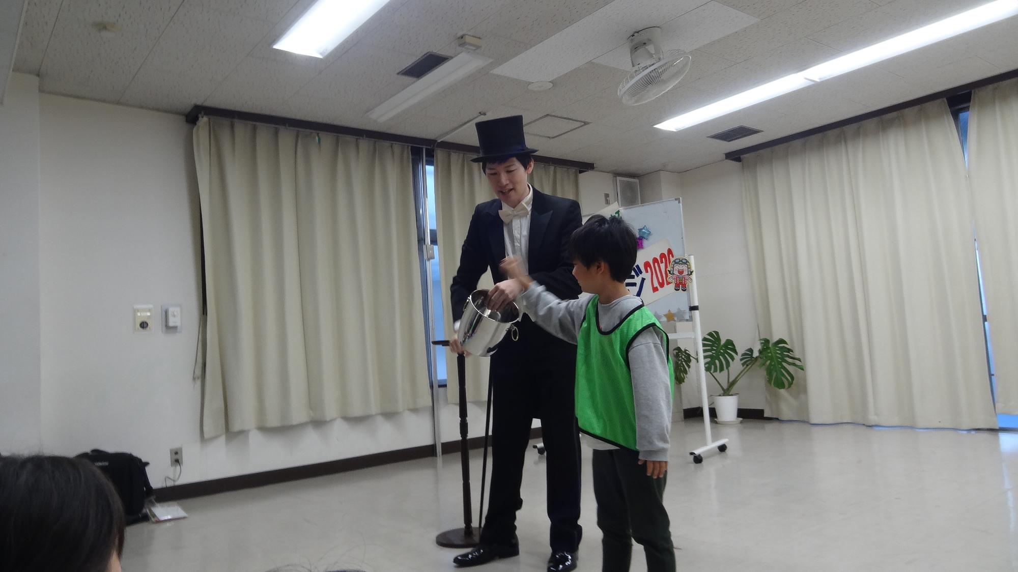 空中をつかんでバケツに入れたらお金の音が鳴ってお金が増えるマジックに小学生1名が参加している様子