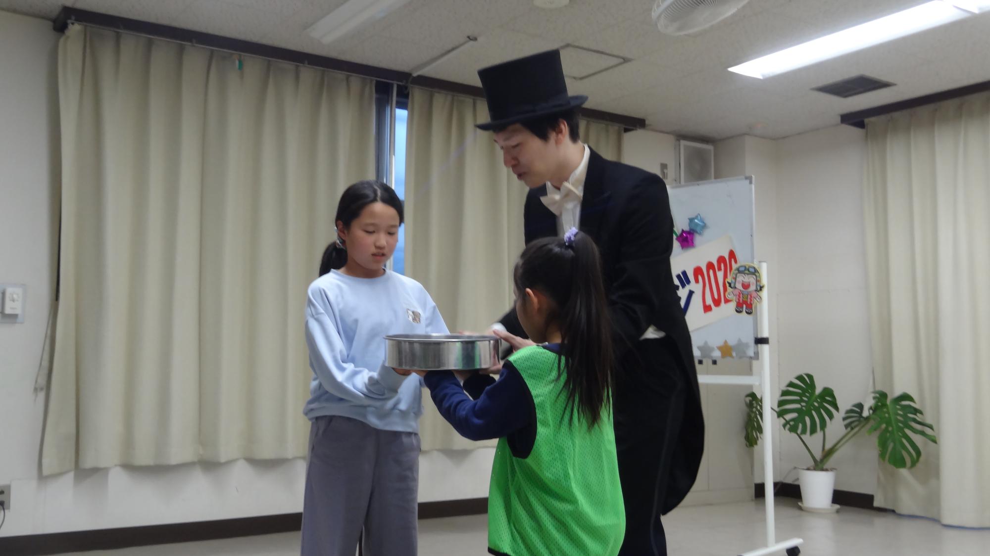 二人の児童が銀色の器を持っている様子。その間にマジシャンがいる。