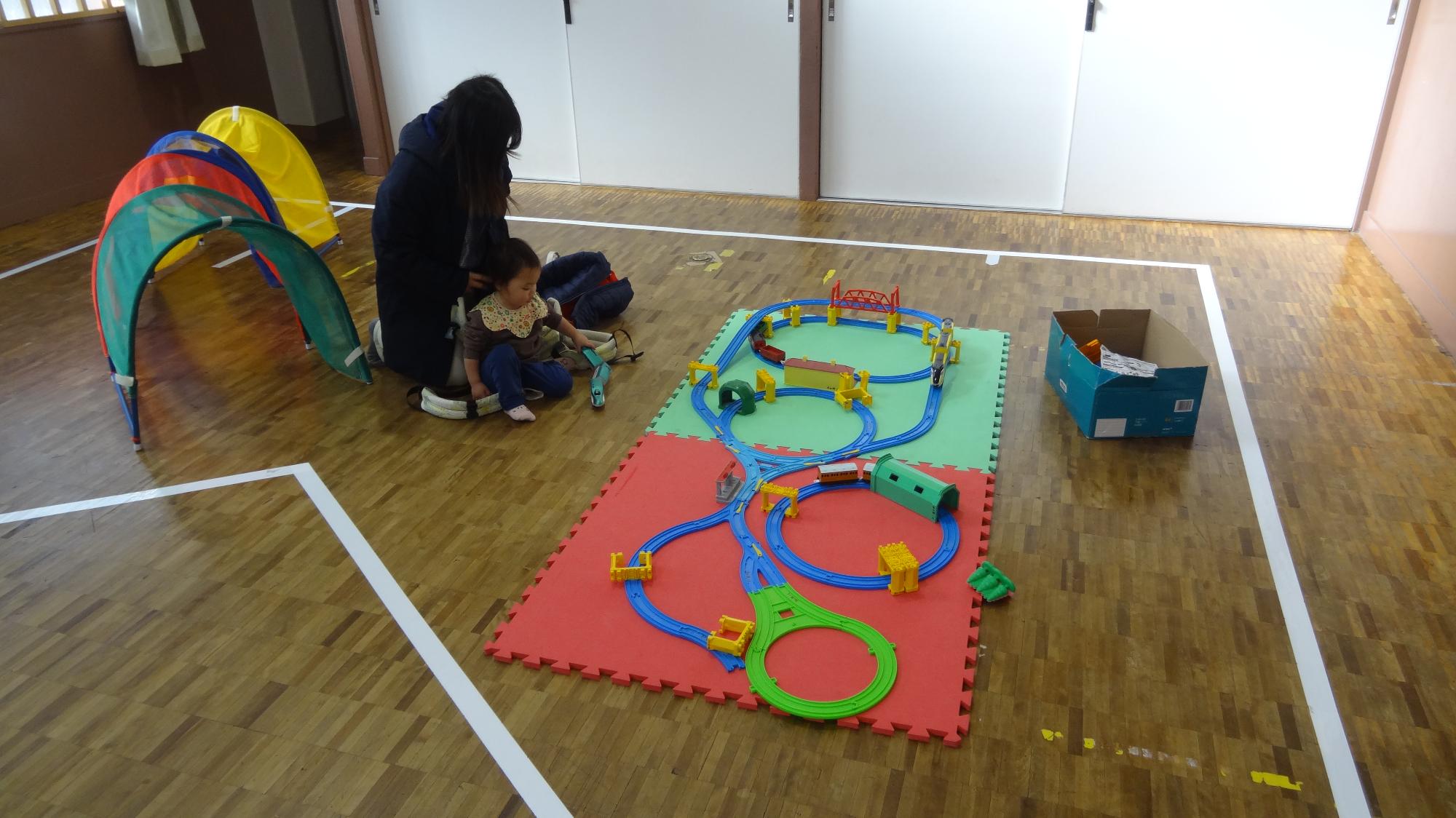 プラレールで保護者の膝の上に座っている幼児が遊んでいる様子。その後ろには、幼児用のトンネルがある。