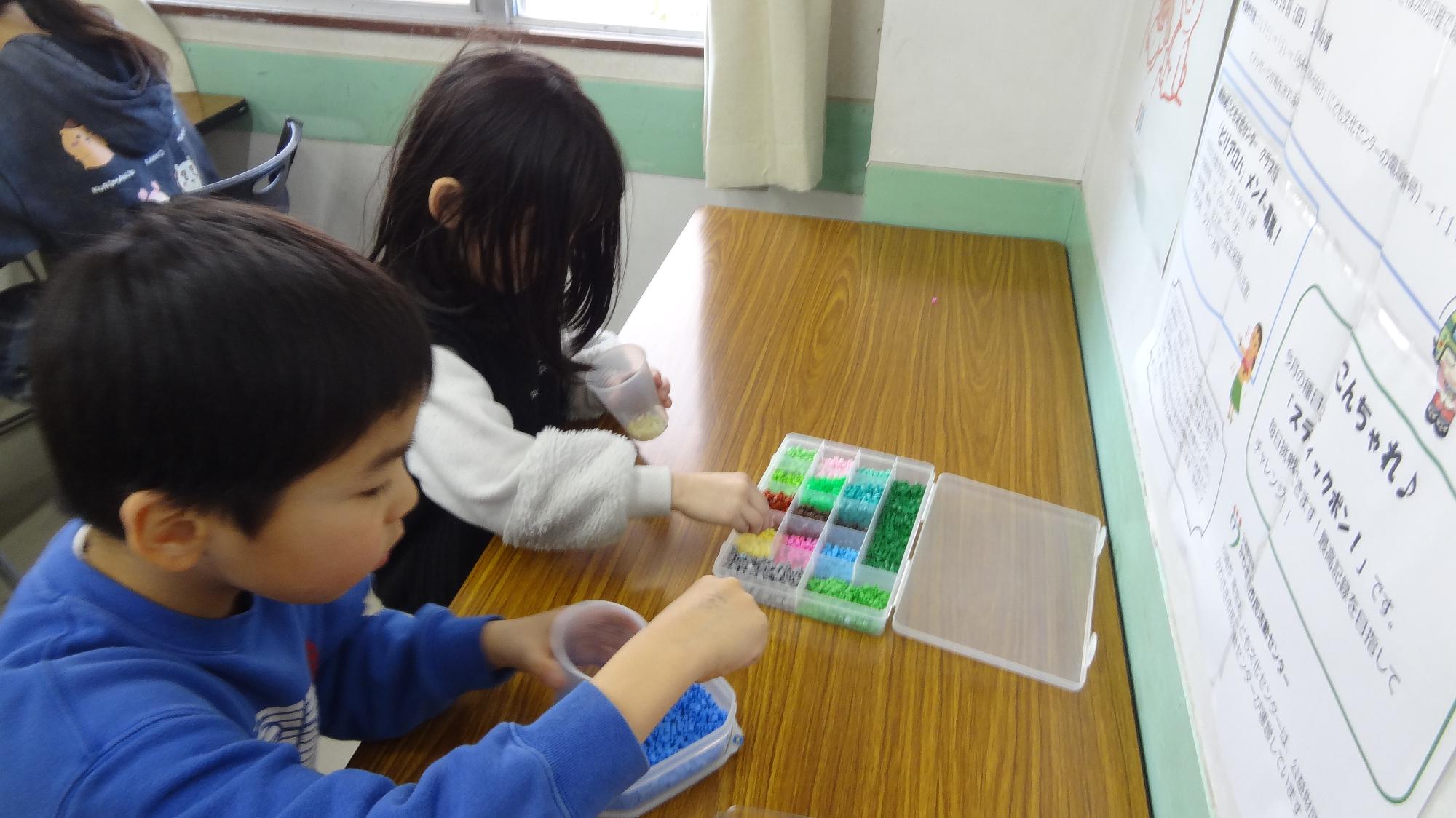 小学生二人が色の着いたビーズをケースの中から選んでいる様子