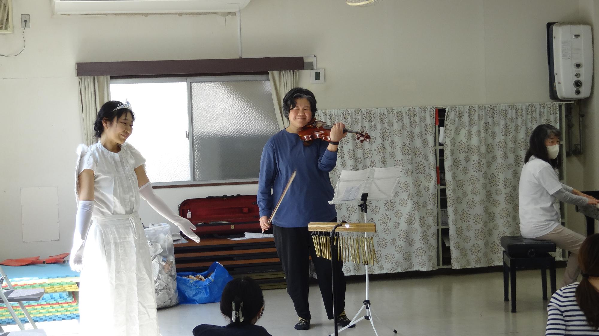 音楽イベントの様子。左の女性は羽の付いた衣装を着ている。真ん中の女性はバイオリンを弾いている。右の女性はピアノを弾いている。