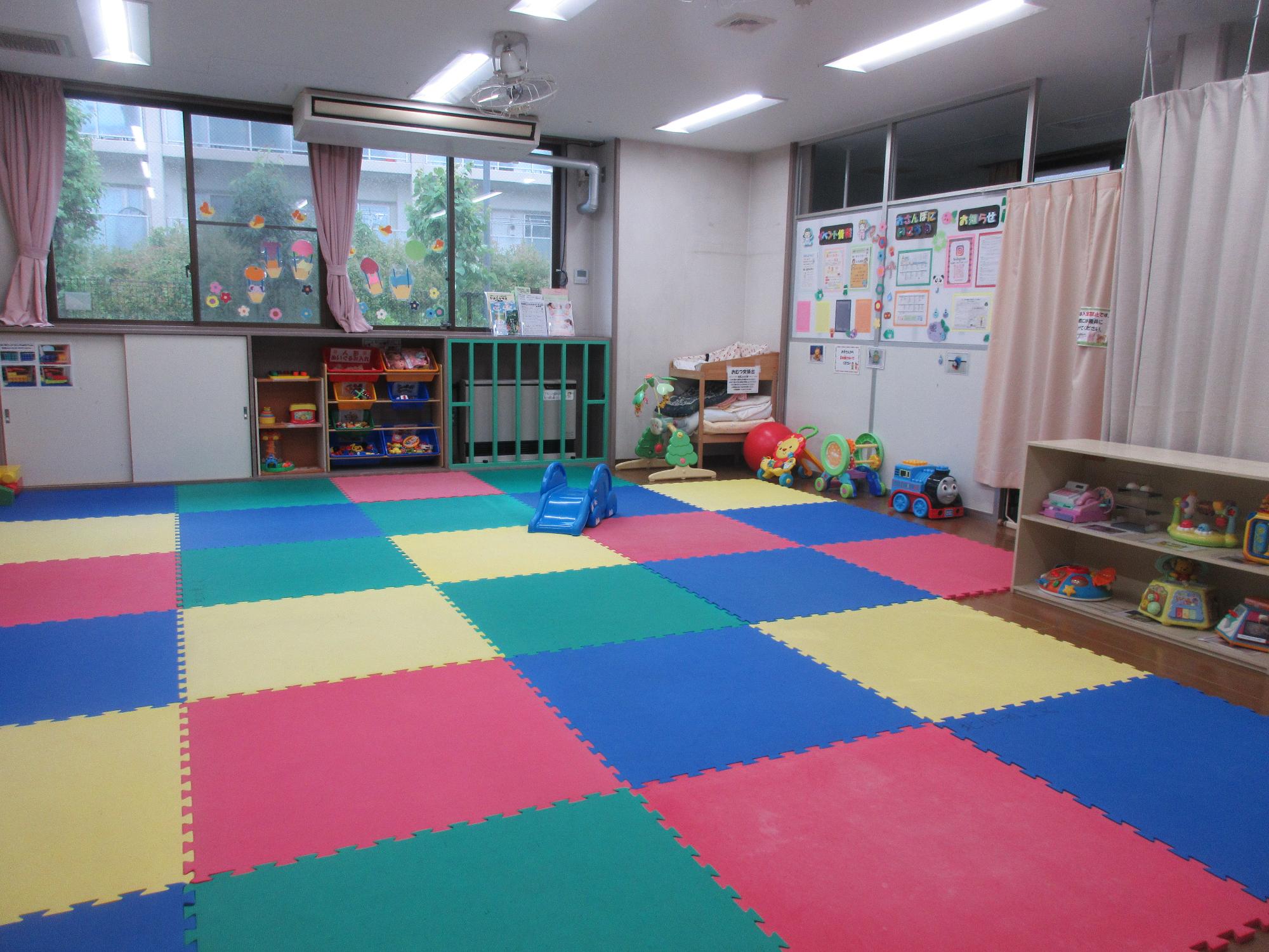 カラフルなマットが敷かれ、おもちゃが置かれた棚、滑り台や遊具が置かれた幼児室の写真