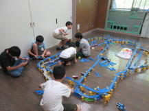 部屋いっぱいに二階建てのプラレールのコースを作っている6人の男の子たちの写真