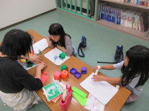 先生が折り方を教えているのを見ている、机を挟んで向い合せに座っている二人の女の子の写真