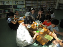 うちわの折り方を教える先生を見ながら一緒に折り紙を折っている子供たちの写真