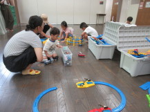 白い箱に沢山入ったレールを並べる3人の子供と一緒にしゃがんで見守る大人たちの写真