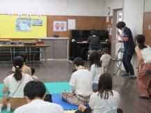 壁際に設置されているピアノを弾く女の子と中央に敷かれたマットに座り演奏を聞く幼児親子の写真