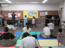 講師の女性と2人の女の子がピアノの演奏に合わせ両手を上げ手遊びをする様子を真似している親子連れの写真