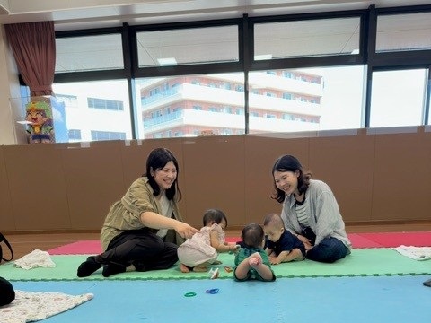 カラフルなマットの上で談笑する乳児親子の写真