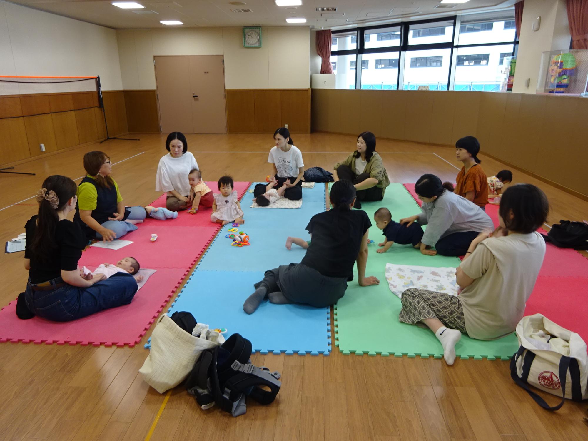 カラフルなマットの上で保育士の話を聞いている8組の乳児親子の写真