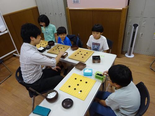 碁盤の前で講師の説明を聞いている4人の小学生の写真