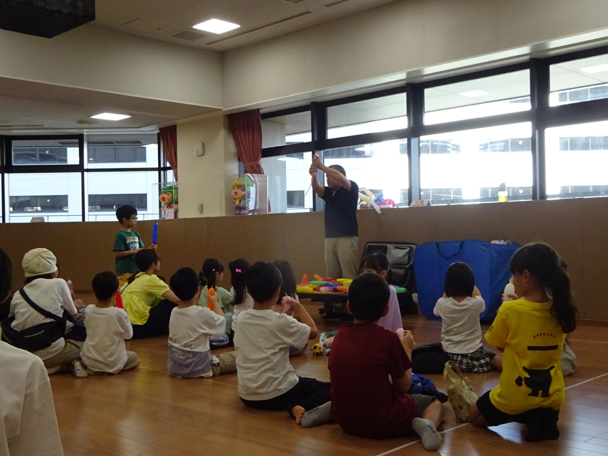 バルーンアート講師の説明を聞いている大勢の子どもたちの写真