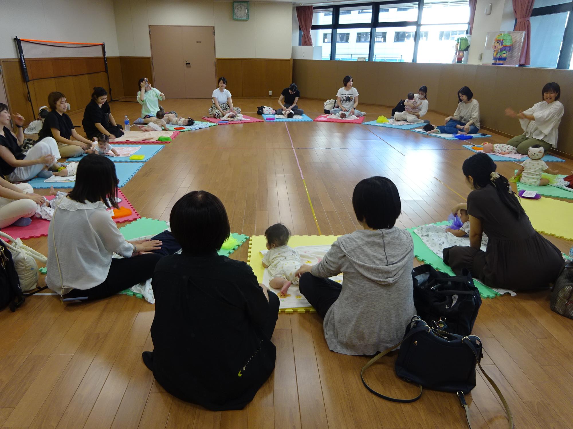 円形に並んでカラフルなマットの上でベビーマッサージをしている15組の親子の写真