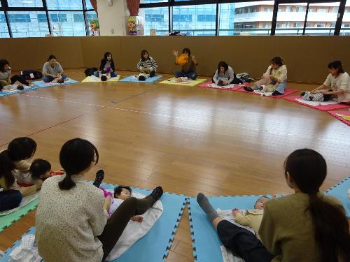 円形に並んでカラフルなマットの上でベビーマッサージをしている17組の親子の写真
