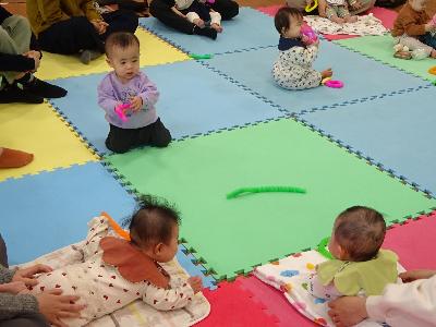 カラフルなマットの上で寝転んだり遊んだりしている乳幼児たちの写真
