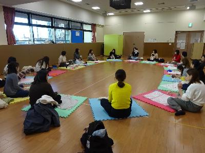 円形に並んでカラフルなマットの上で自己紹介をしている16組の親子の写真