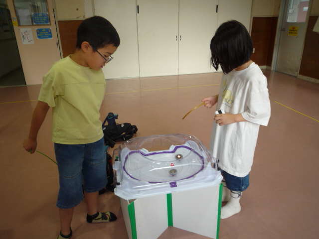 小学生の男の子と女の子がベイブレードの勝負の行方を見守ってる写真