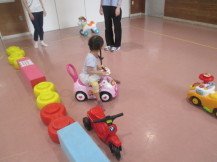 ピンク色の床の室内で、大人の方に見守られながらピンク色の玩具の車に乗って遊んでいる女の子の写真