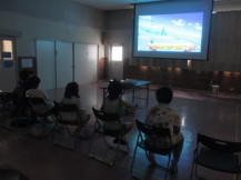 うす暗い室内でスクリーンに映った画面を見ながらゲームをし、横一列に並べられた折りたたみ椅子に座っている子どもたちの写真