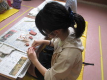 新聞紙の敷かれたテーブルの前でカラーマットの上に座り、茶色の粘土を使って土偶を作っている女の子の写真