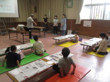 室内の前のほうに立ち話をしている講師の方と、新聞紙の敷かれたテーブルの前でカラーマットの上に座っている子どもたちの写真