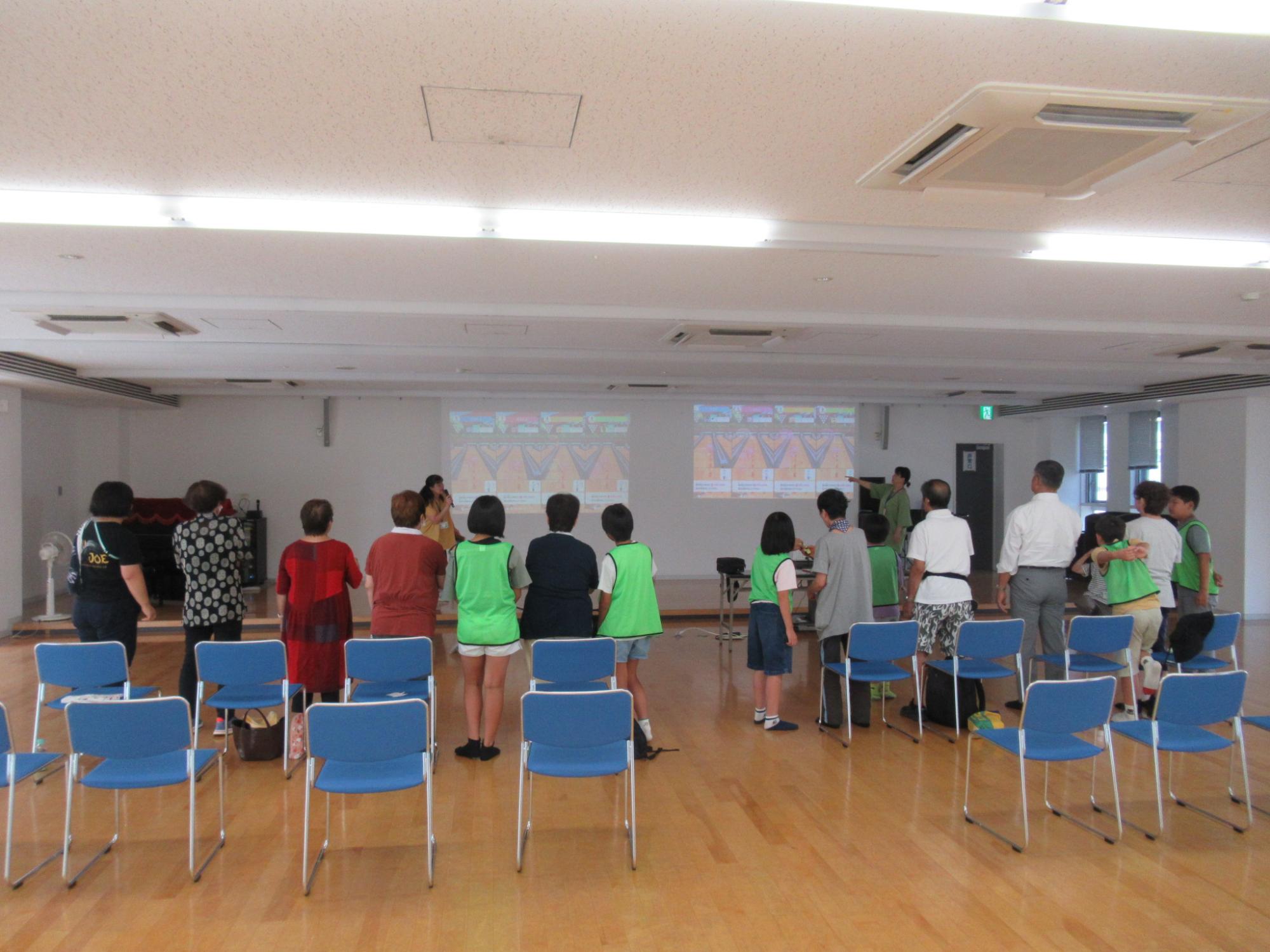 スクリーンに映されたゲーム画面を見ながらボウリングゲームを楽しむ老人と小学生の写真