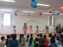 ステージ上でダンスを披露する5人の女の子と座って見ている大勢の子供たちの写真
