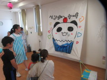 パンダとケーキを合わせた絵が飾られた部屋で絵を鑑賞する親子たちの写真
