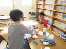 ポストカードづくりに必要な材料や文房具が置かれた長机の席に座っている二人の子供の写真