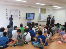 大勢の子供たちが集まる中モニターを使いオンラインでビンゴ大会をしている様子を撮影している写真