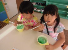 子供が2人並んで座り、メロンのかき氷をストローで口に運びながら食べている様子の写真