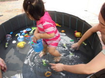 折りたたみプールに浮いているおもちゃをピンクの水着を着た幼児が小さなバケツに集めている様子を写した写真