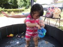 折りたたみプールの中でバケツを覗き込んでいるピンクの水着を着た幼児の写真
