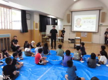 室内に敷かれたブルーシートにたくさんの子供たちが座り前方のスクリーンをみている様子を後方から写した写真