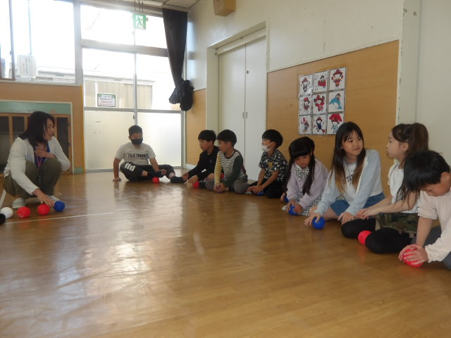 集会室で職員と一緒にボッチャで使うボールを転がして柔らかくしている小学生たちの写真