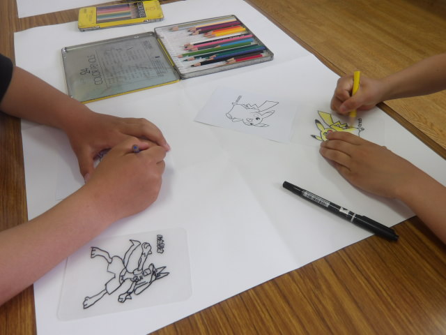 アニメのキャラクターが描かれたプラバンに色鉛筆で色を塗る小学生2人の写真