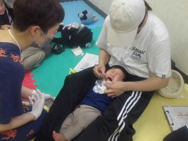 幼児さんに歯磨きをするお母さんと、歯磨きが嫌で泣く幼児さんと、歯磨きの指導をする講師の写真