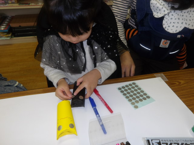 トイレットペーパーの芯に黒の折り紙を貼った芯に丸シールを貼って顔を作る幼児の写真