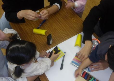 談笑しながら幼児を抱えるお母さん達とトイレットペーパーの芯に黒の折り紙を貼った芯に丸シールやペンで顔を作る幼児達の写真