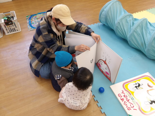 大きな絵本を読み聞かせするお母さんと読み聞かせを聞く幼児2人の写真
