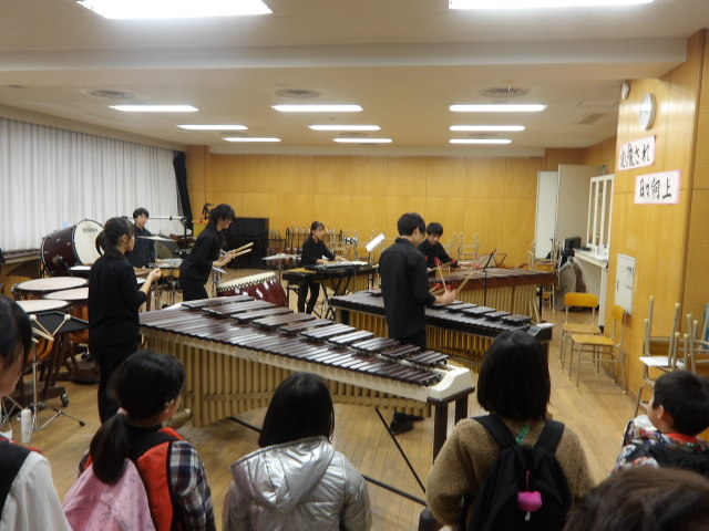 音楽室で高校生達の練習を見る小学生達の写真