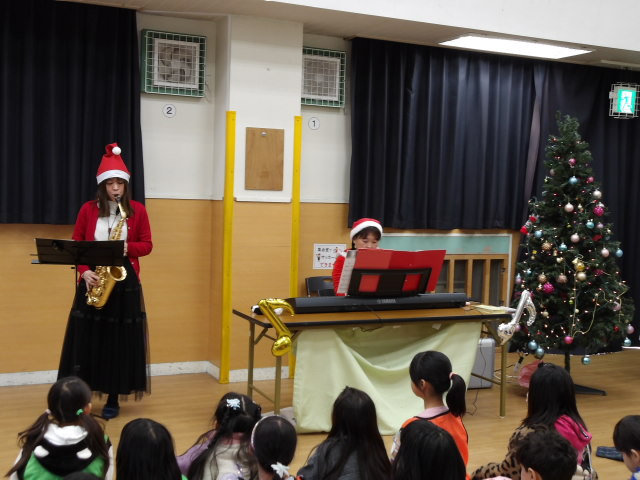 サックスを演奏する演奏者とピアノを伴奏する講師と演奏を聴く小学生達の写真