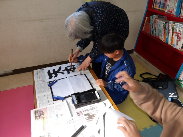 書道の先生と一緒に筆を持ちながら、半紙に字を書く小学生の写真